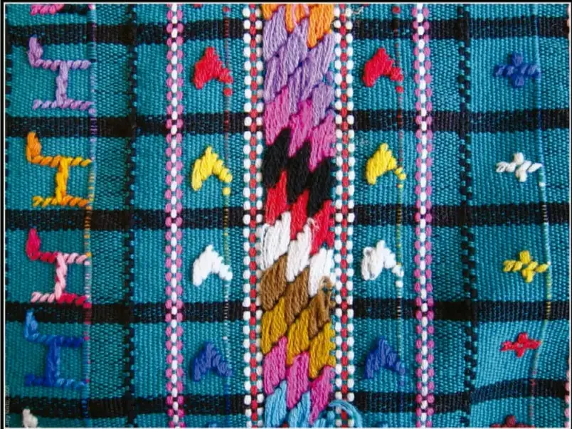 Fotografía de una tela tejida a cuadros en azul turquesa y negro, con bordados de figuras humanas en posturas diversas, flores y animales en rojo, amarillo, blanco y azul; al centro una franja vertical con un tejido tipo petate o plumas de colores rosa, morado, naranja y verde.