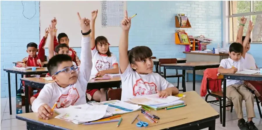 Fotografía de un salón de clases con niños y niñas de uniforme blanco sentados en sus pupitres, varios levantan la mano con el dedo índice para participar; hay libros, crayones y tijeras sobre los pupitres.