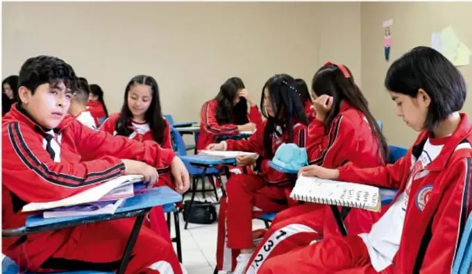 Fotografía de estudiantes de telesecundaria con uniforme rojo trabajando en equipo con sus cuadernos en el aula.