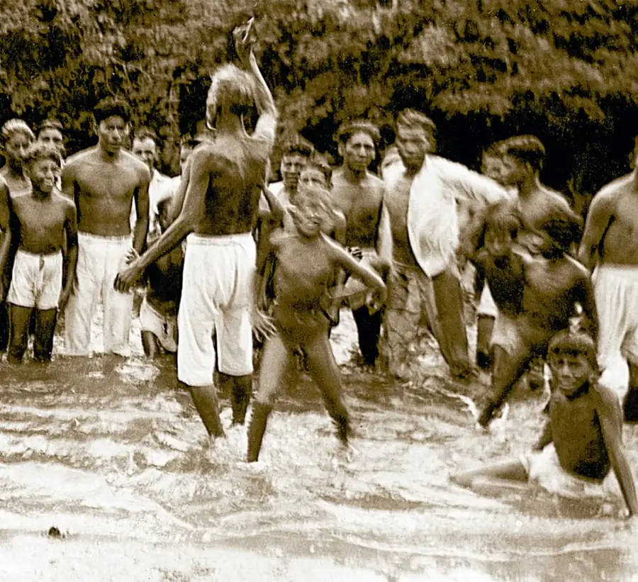 Fotografía histórica en tono sepia: grupo de niños y jóvenes, en traje de baño o pantalones blancos, jugando y chapoteando en el agua (río o mar), con árboles y vegetación densa al fondo. Ilustra la vida cotidiana y el recreo de las comunidades costeras.