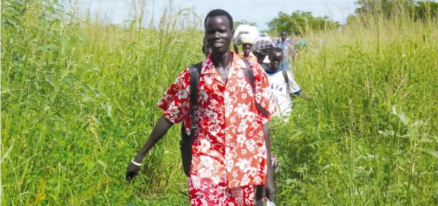 Grupo de personas migrantes caminando entre pastizales altos; al frente una mujer con ropa roja de flores y mochila, seguida de otras personas.