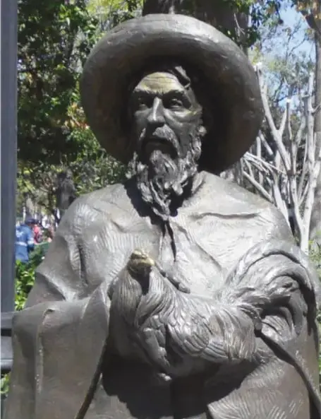 Estatua de bronce del gallero: hombre de sombrero ancho, barba y manta, que carga un gallo entre sus brazos.