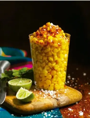 Vaso con granos de maíz amarillo cocido, acompañado de limones y una cuchara sobre una tabla de madera.