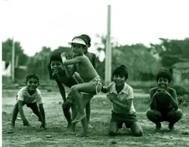 Fotografía en blanco y negro (impresa en tinta verde) de seis niños descalzos en poses de juego de béisbol en un campo; al centro el lanzador y a un lado un niño en cuclillas con la pelota; al fondo árboles y un poste.