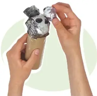 Manos colocando pequeñas piezas de papel cubiertas de cinta sobre la cabeza del alebrije (tubo con tapa y bola de papel).