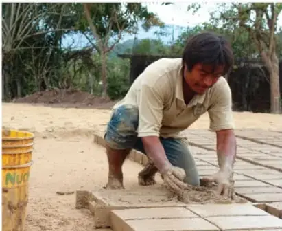 Hombre joven que elabora manualmente piezas de piso (adoquines) con mezcla de arena y cemento, trabajando de rodillas.