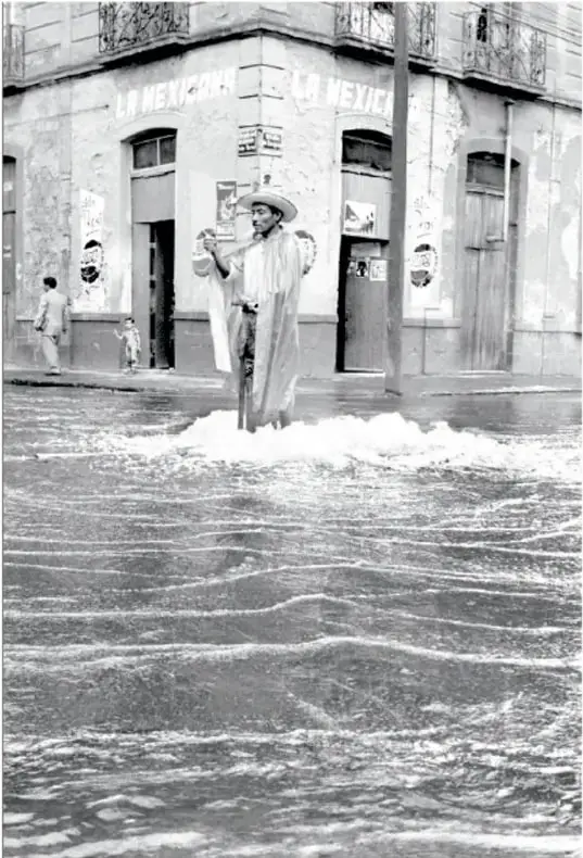 Fotografía en blanco y negro de una calle inundada. Un hombre con sombrero y un lienzo blanco camina entre el agua. Al fondo, una vecindad con la palabra 'MÉXICO' pintada. Título: 'Tlaloc, ca. 1960, Héctor García (1923-2012)'.