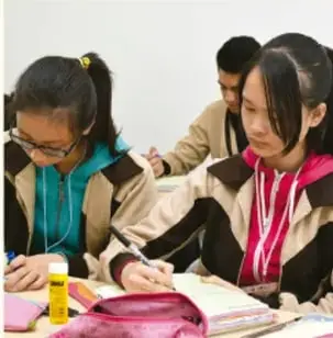 Fotografía de tres estudiantes trabajando en equipo: una joven escribe en un cuaderno sobre la mesa mientras sus compañeros revisan materiales de colores.