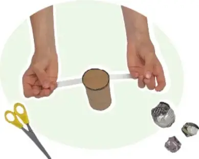 Manos tensando una tira de cinta adhesiva sobre un tubo de cartón; cerca, tijeras amarillas y bolas de papel arrugado.