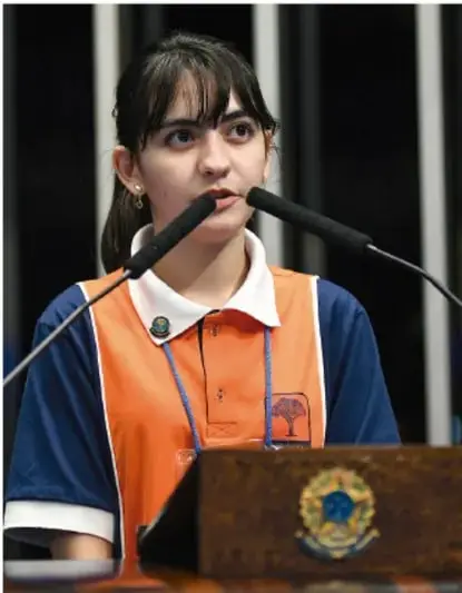 Fotografía de una adolescente hablando frente a un micrófono, con uniforme escolar, en un espacio de participación o foro.