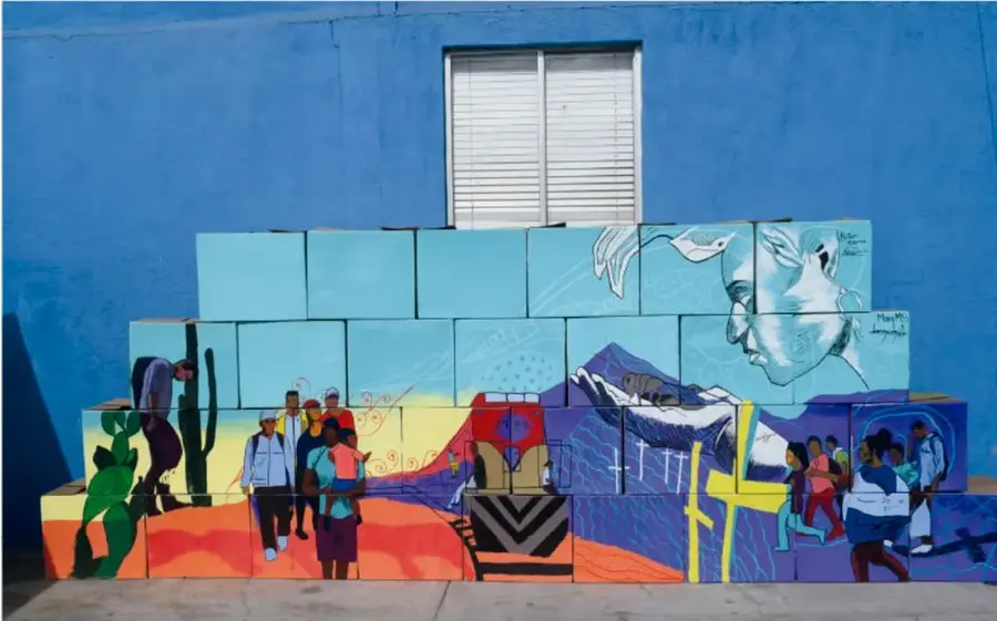 Mural pintado en bloques de madera frente a un muro azul: personas caminando con bultos, cactus, un toro, montañas azules y una paloma blanca; representa a personas migrantes.