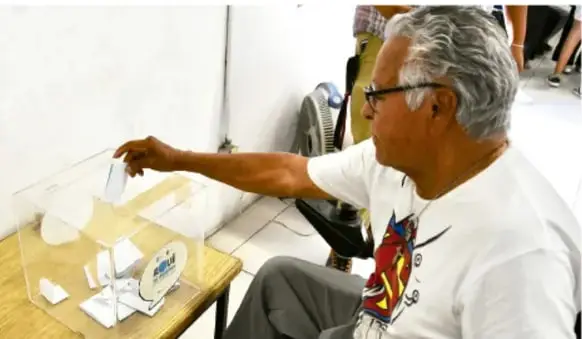 Fotografía de una persona adulta que deposita un papel (su voto) dentro de una urna transparente durante una jornada electoral.