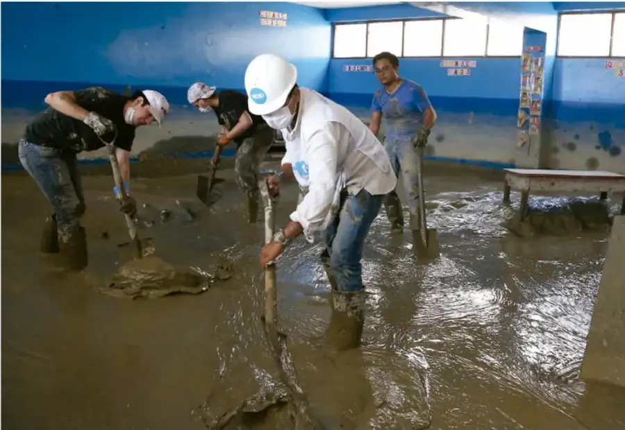Fotografía de varias personas con botas, guantes y cubrebocas paleando lodo y agua dentro de un salón de clases inundado, con paredes azules y bancas escolares.