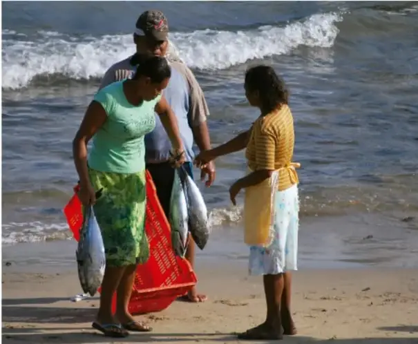Fotografía de tres personas en la playa junto al mar, cargando peces y una red roja, ilustrando actividades comunitarias de pueblos costeros.