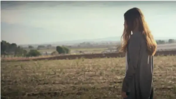 Fotograma del cortometraje: una joven de espaldas con cabello largo, de pie en un campo abierto bajo un cielo nublado.