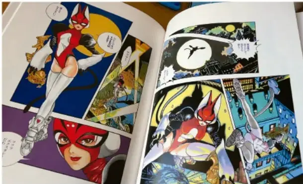 Fotografía de un cómic o manga abierto que muestra viñetas de una superheroína con máscara y traje rojo, y un personaje con máscara de zorro volando sobre una ciudad nocturna con luna llena.