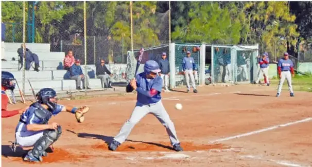 Partido de béisbol en el patio de una escuela: el bateador en posición de golpear, el receptor acuartelado con guante, un umpire y jugadores de ambos equipos en posición; hay un balón blanco en pleno vuelo hacia el bate.