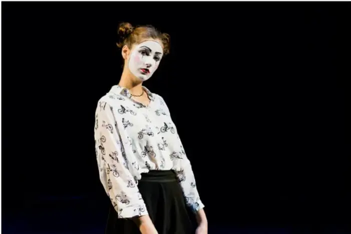 Fotografía de una actriz con maquillaje blanco de estilo mimo o payaso, caminando sobre un escenario con fondo negro, iluminada por luz de teatro.