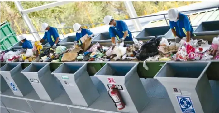 Trabajadores con uniforme azul y casco blanco separan residuos en una banda transportadora de una planta de reciclaje, con contenedores de colores al frente.