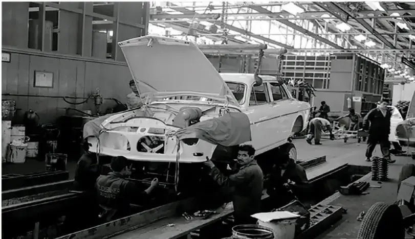 Fotografía en blanco y negro de una línea de ensamblaje automotriz. Varios trabajadores con overol manipulan la carrocería de un automóvil clásico dentro de una nave industrial con estructura metálica y grandes ventanales. Se observan herramientas, neumáticos y cajas en el piso.