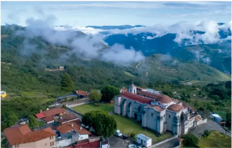 Fotografía aérea de Santa Catarina Lachatao, Oaxaca, con un templo colonial rodeado de casas de techos rojos, cerros verdes y nubes bajas; crédito: Fulvio Eccardi.
