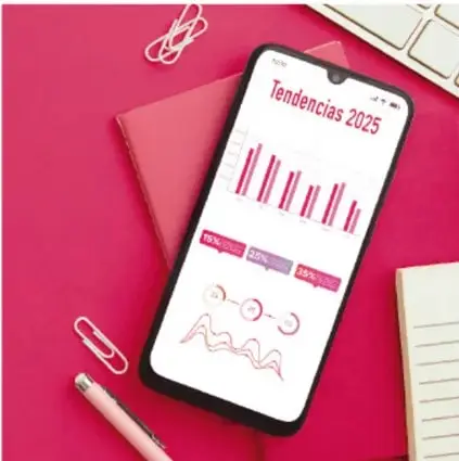 Fotografía de un escritorio rosa con un teléfono móvil que muestra una gráfica de barras titulada 'Tendencias 2025', junto a cuaderno, clips y bolígrafo.