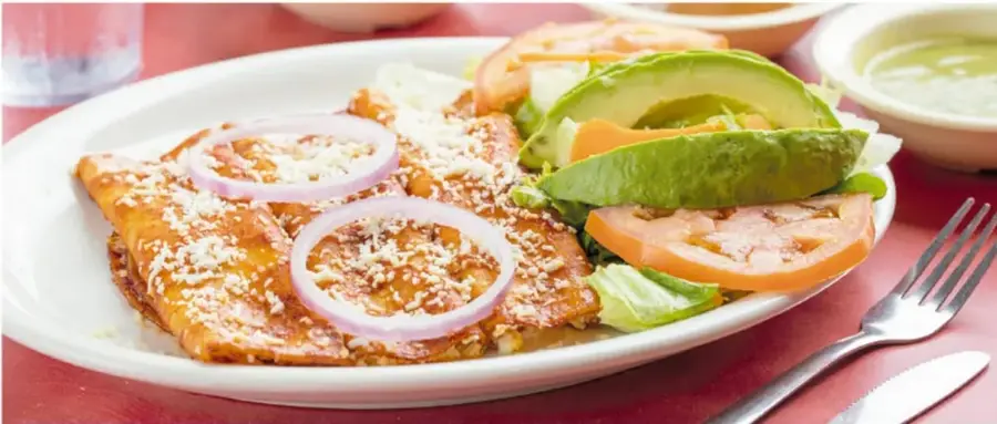 Fotografía de un plato de enchiladas potosinas rojas con queso espolvoreado y aros de cebolla, acompañadas de rebanadas de aguacate, jitomate y lechuga, sobre mantel rojo.