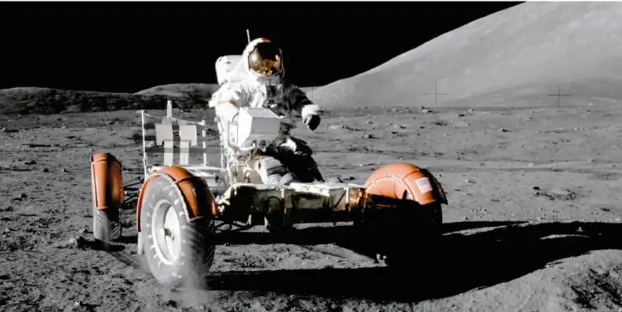 Fotografía en blanco y negro de un astronauta conduciendo un vehículo lunar (rover) sobre la superficie de la Luna, con un paisaje árido y montañoso de fondo.