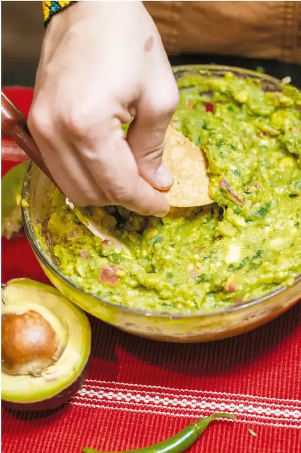 Fotografía de una mano metiendo un totopo (nachos) a un tazón de vidrio con guacamole recién hecho; junto al tazón hay medio aguacate con hueso y un chile serrano sobre un mantel rojo tejido.