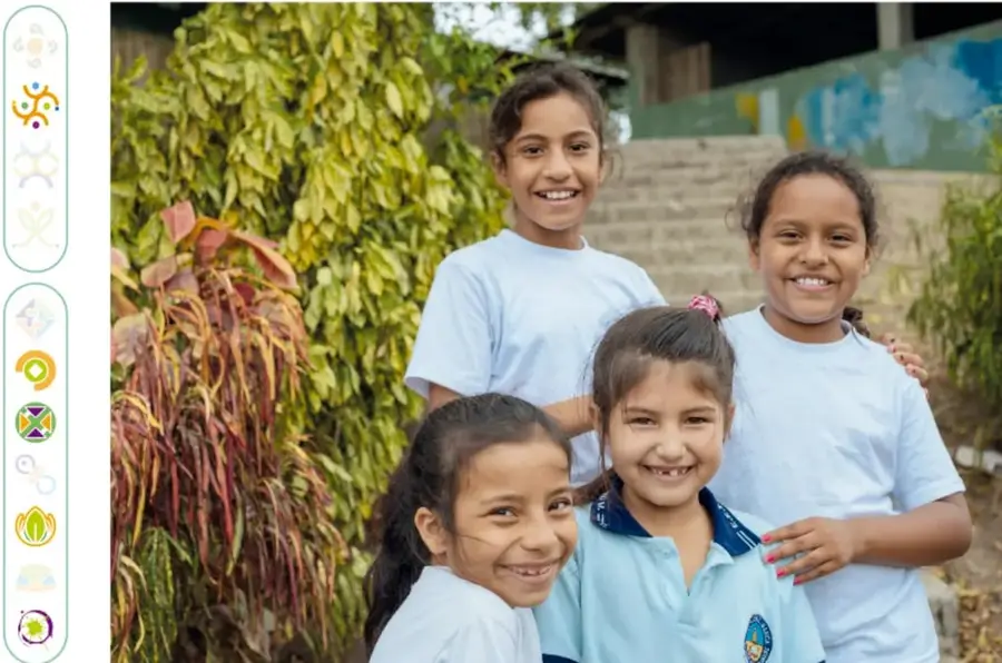 Fotografía de cuatro niñas sonrientes de camiseta blanca, juntas frente a plantas ornamentales y una escalera de piedra, al aire libre.