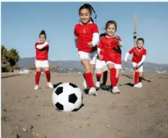 Fotografía de niñas y niños con uniforme deportivo rojo jugando futbol en una cancha de tierra, con un balón blanco y negro al frente.