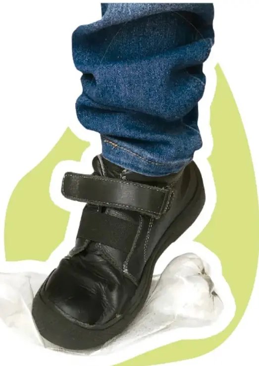 Fotografía de un pie con zapato escolar de piel y pantalón de mezclilla aplastando una botella plástica PET transparente sobre un fondo verde difuso.