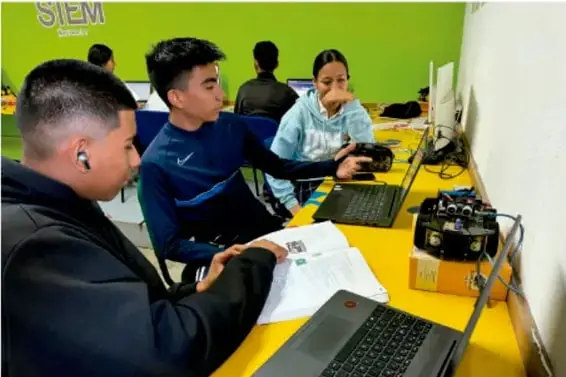 Fotografía de cuatro estudiantes de Telesecundaria trabajando en equipo alrededor de una mesa con laptops, un cuaderno abierto y equipo electrónico, en un aula con computadoras.