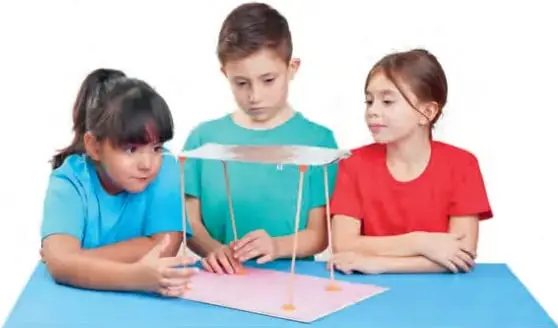 Tres estudiantes presentan una maqueta sobre una base rosada: una niña señala y explica, mientras los otros dos observan y sostienen el trabajo.