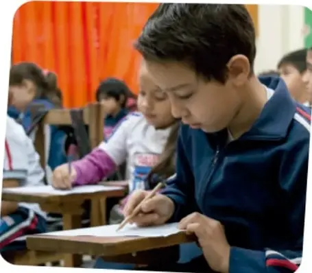 Niño sentado en su pupitre escribe en una hoja mientras otros compañeros trabajan en el salón.