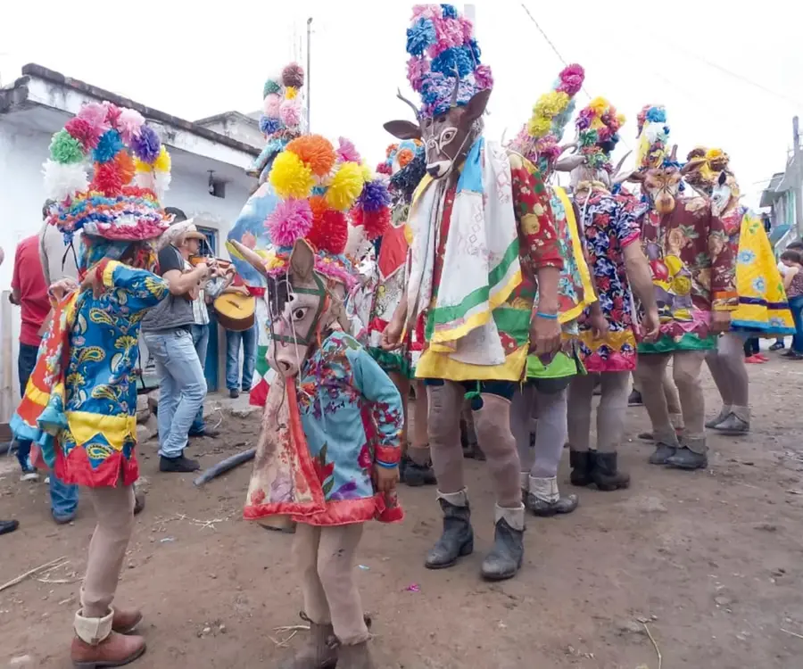 Carnaval en un pueblo: personas con máscaras de venado y burro y enormes tocados de pompones y flores de papel de colores; al fondo, músicos con guitarra.