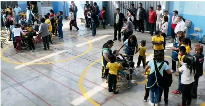 Fotografía de un simulacro escolar en un patio. Se observan estudiantes y adultos organizados en filas, algunos con chalecos de protección civil, y una persona en silla de ruedas siendo evacuada. El piso tiene marcas amarillas y rojas que indican rutas o zonas de seguridad.
