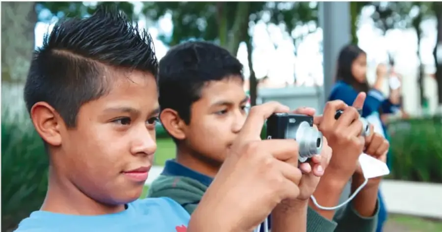 Fotografía de dos adolescentes en un parque; uno sostiene una cámara compacta mirando su pantalla y otro revisa un teléfono celular; al fondo hay árboles y una joven de pie.
