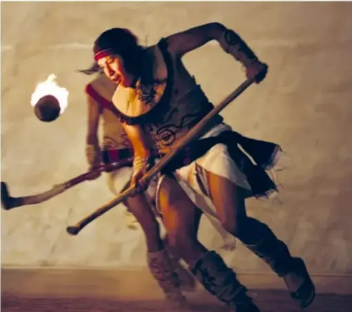Fotografía de dos jugadoras de pelota purépecha en movimiento, con bastones y una pelota encendida, vistiendo ropa tradicional.