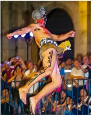 Fotografía de una persona con vestimenta tradicional y pintura corporal saltando en un evento comunitario frente a una multitud, en un acto festivo que puede vincularse con juegos o tradiciones con pelota.