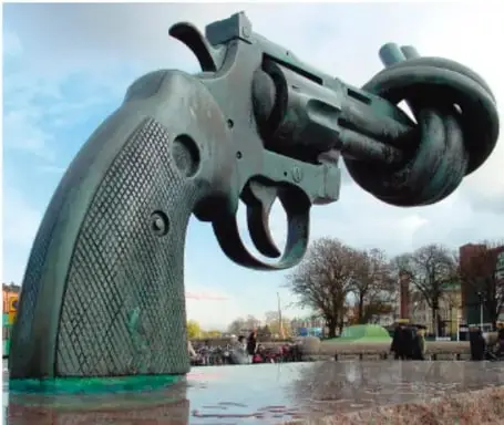 Escultura de bronce de un revólver con el cañón anudado, conocida como 'Non Violence' (obra de Carl Fredrik Reuterswärd), ubicada al aire libre junto a un estanque y árboles. Simboliza el rechazo a la violencia.