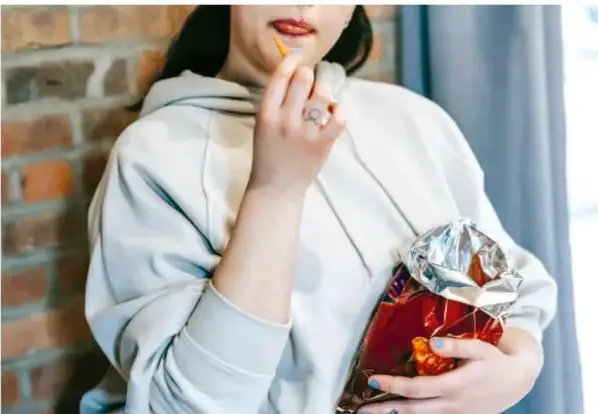Fotografía de una persona joven comiendo una fritura de una bolsa, con ladrillo de fondo.