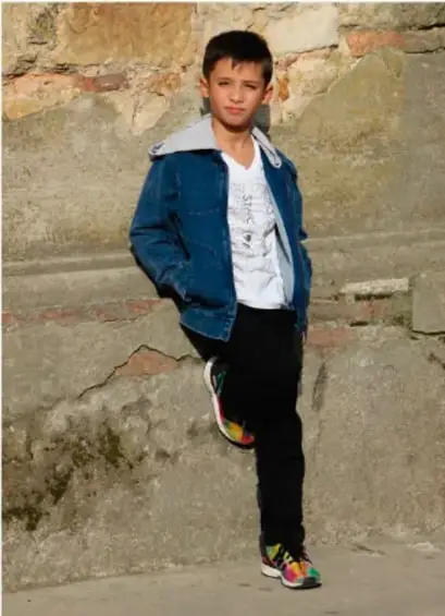 Fotografía de un niño de primaria con chamarra azul y pantalón negro, recargado en un muro de piedra.