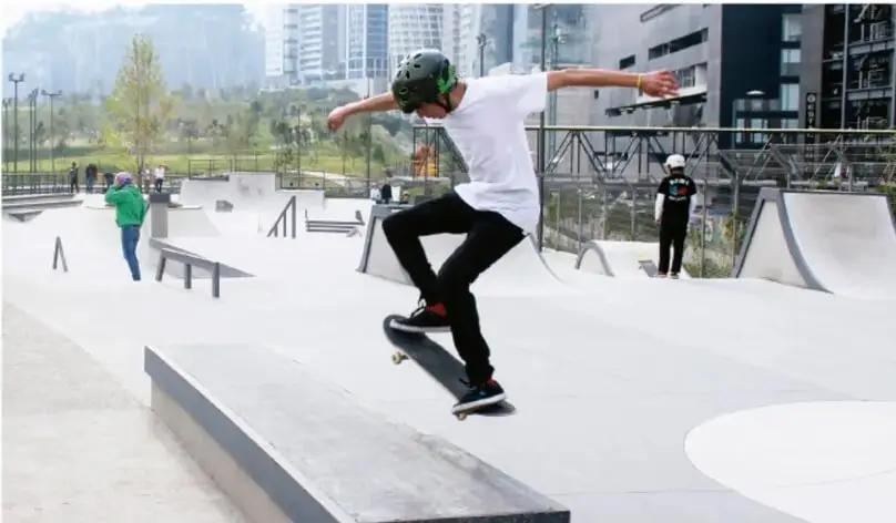 Fotografía de un joven skatista con casco realizando un truco sobre una plataforma baja en un skatepark urbano; al fondo hay rampas, otras personas y edificios de la ciudad.