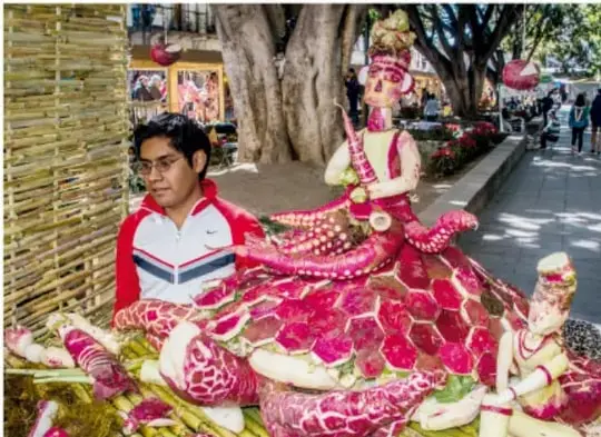 Persona joven junto a una figura artesanal vestida con traje colorido y bordado, en una calle con árboles, en una muestra cultural o festividad tradicional.