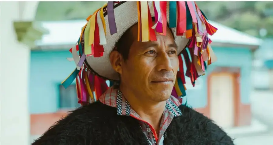 Fotografía de un hombre indígena con sombrero adornado con cintas de colores, posiblemente relacionada con la tradición cultural descrita.