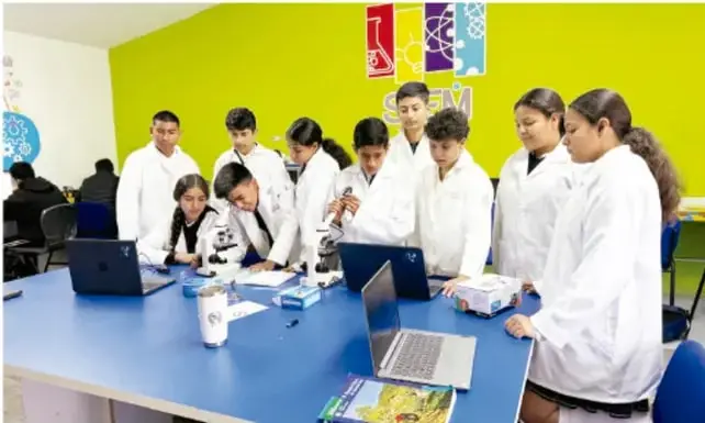 Fotografía de estudiantes con batas blancas en un laboratorio escolar, trabajando con microscopios, laptops y materiales sobre una mesa azul; pared amarilla con logotipos de ciencia.