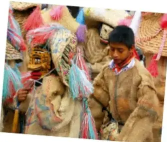 Grupo de niños y adultos con trajes de danza tradicional: máscaras, sombreros adornados y ropa típica, participando en una fiesta comunitaria.