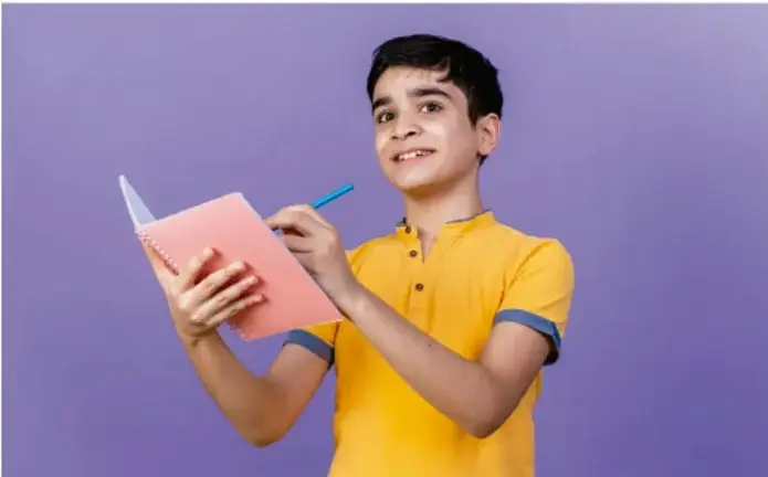 Fotografía de un estudiante de secundaria con camiseta amarilla sobre fondo morado, sonriendo mientras sostiene un cuaderno rosado y un lápiz, en actitud de escribir o reflexionar.