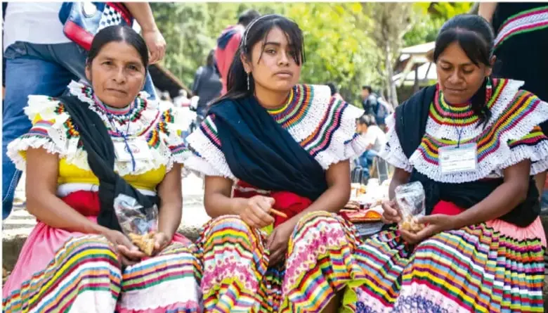 Fotografía de tres mujeres indígenas de_traje tradicional colorido (huipil y falda bordada), sentadas en un espacio al aire libre, participando en una actividad comunitaria; se relaciona con la identidad cultural y la comunidad.